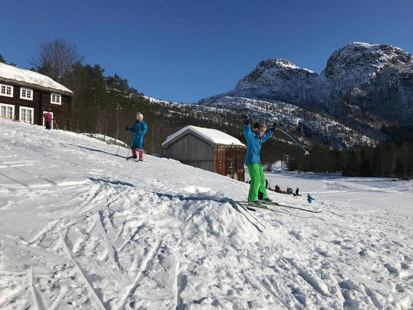 Et barn hopper på et skihopp ved en gammel hytte med store fjell i bakgrunnen.