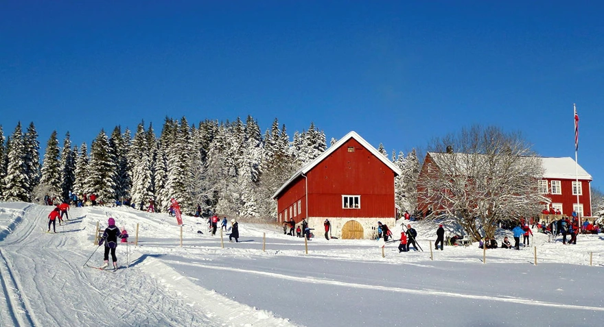 En stor rød hytte og en rød låve i utkanten av en skog, vinter og masse snø på trærne. Mennesker går i oppkjørte skispor foran hytta.