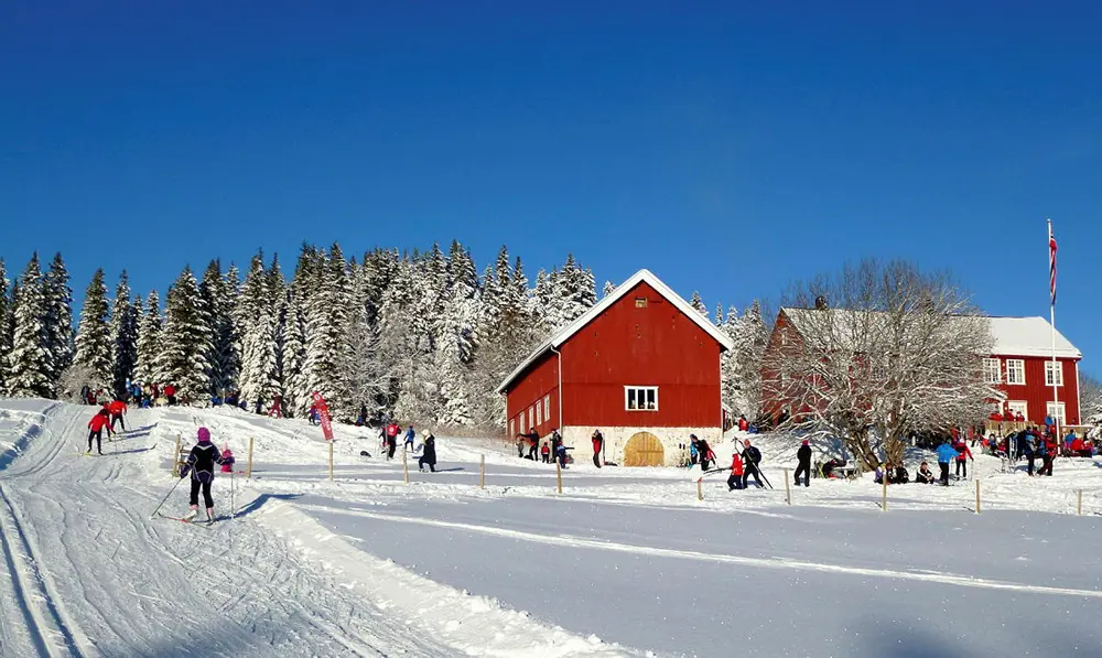 En stor rød hytte og en rød låve i utkanten av en skog, vinter og masse snø på trærne. Mennesker går i oppkjørte skispor foran hytta.