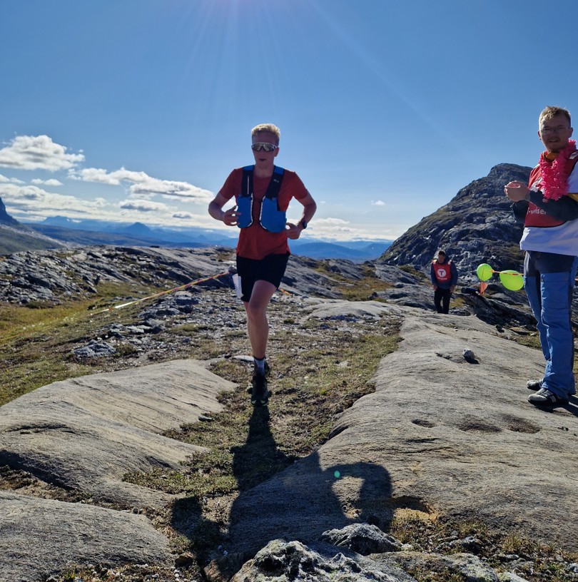 En fjelløper løper over et nakent berg. Han har startnummer på. Det står noen folk på siden og heier på ham.
