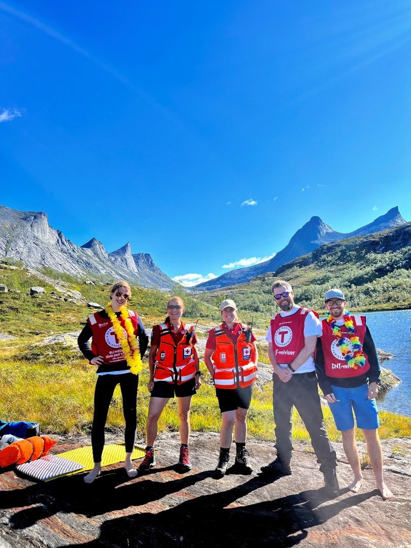 En gjeng ungdommer står på rekke og rad foran et vann. De er på fjellet. Det er strålende sol og de smiler.