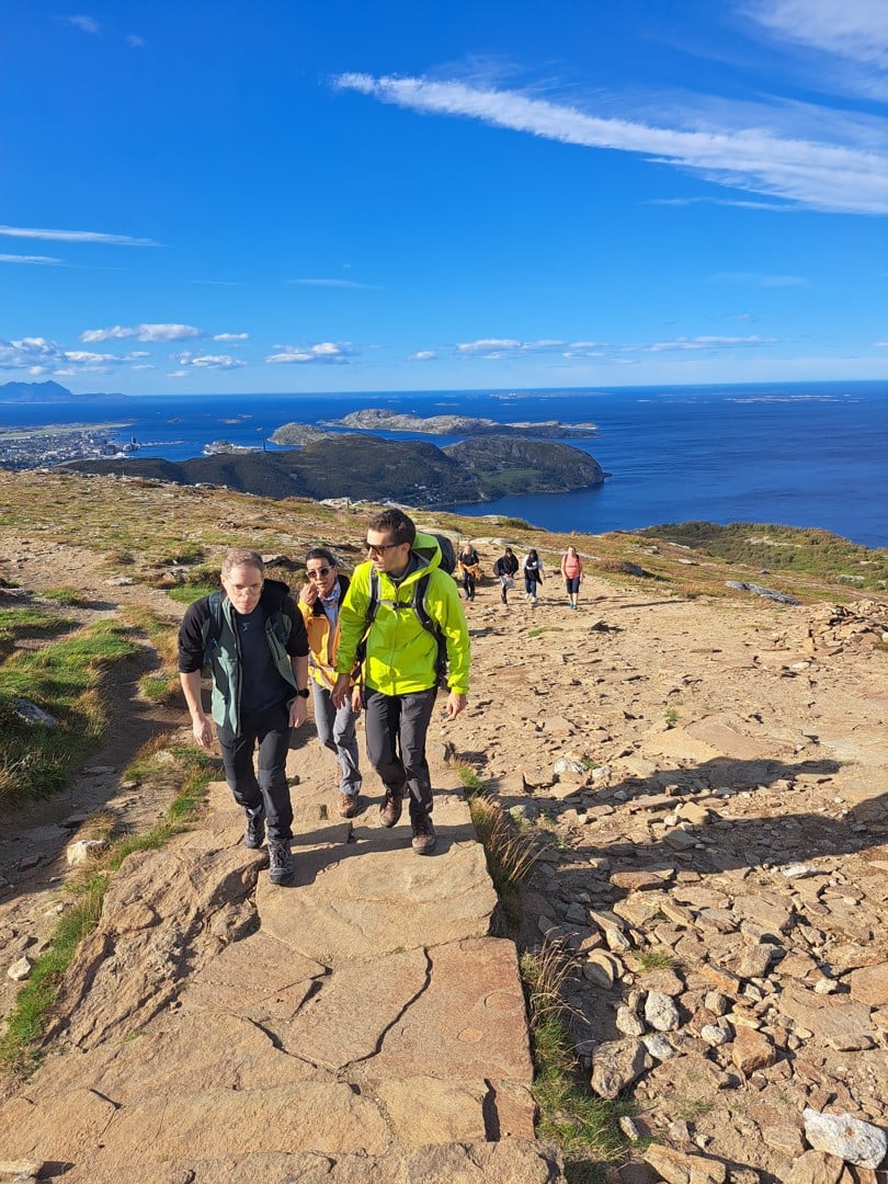 En gjeng folk med friluftsklær går en steintrapp opp et fjell. Bak ser man by og et hav med øyer og fjell. Det er fint vær og blå himmel.