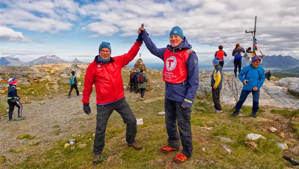 En mann i rød jakke gjør "high five" med en mann i blå jakke og rød vest. Begge har lue på. Det står på toppen av et fjell med utsikt. Det går flere folk i bakgrunnen.