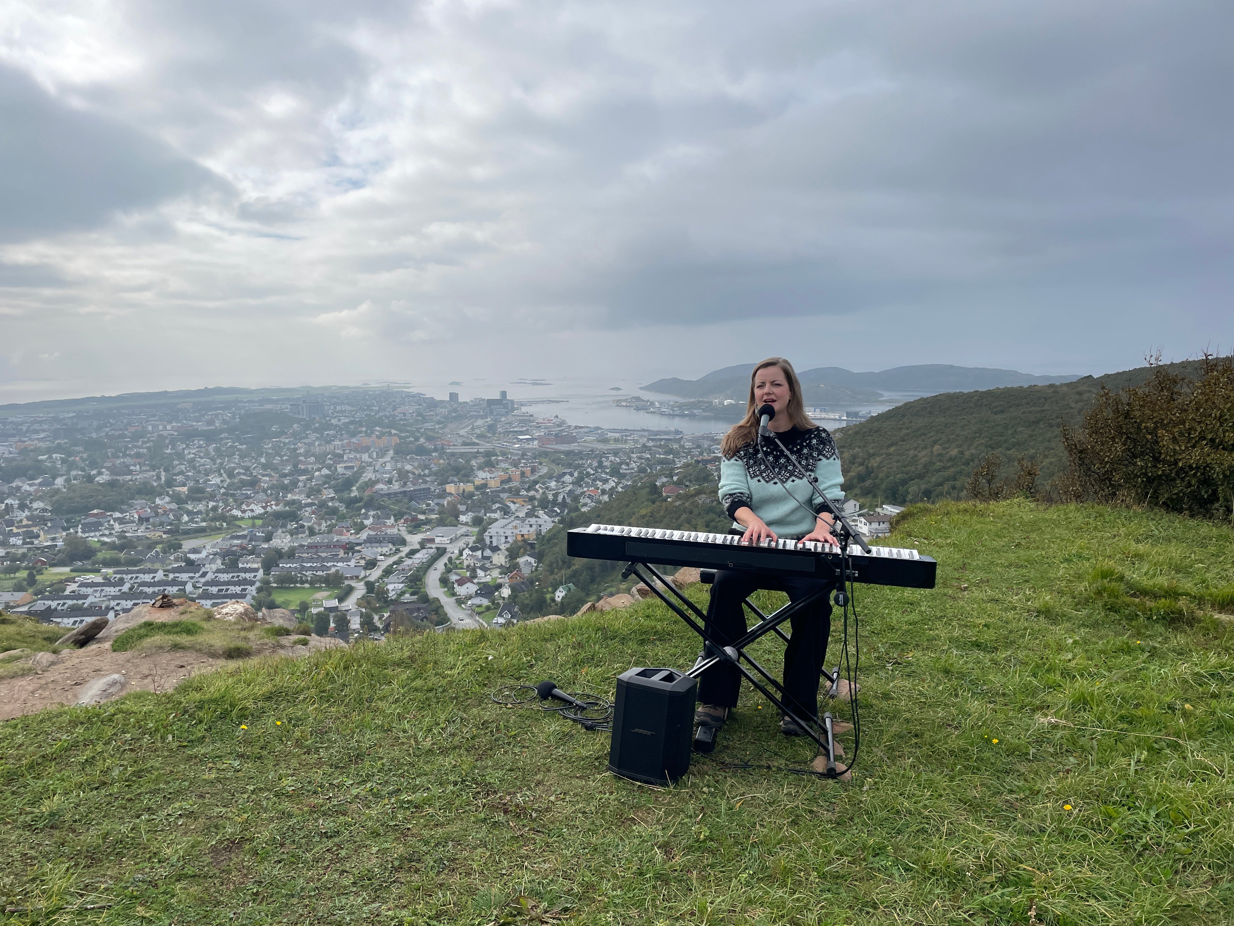 En dame med blå strikkegenser sitter foran et keyboard og en mikrofon og synger og spiller. Hun sitter på kanten av en fjelltopp, og det er utsikt ned mot bebyggelse nedenfor.