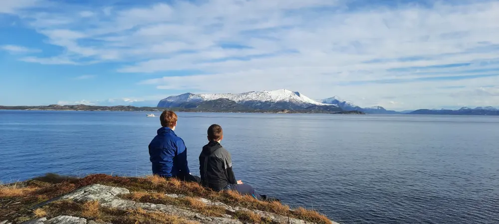 To personer sitter på berget på Sandvikstien i Kristiansund med ryggen til. De har utsikt mot Tustnafjella