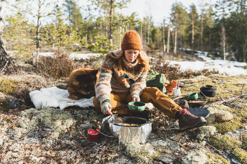 Kvinne sitter på bakken i skogen og lager mat på primus. Ved siden av henne ligger en brun hund.