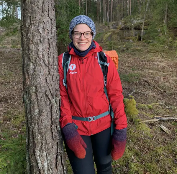 Smilende kvinne i rød DNT-jakke som lener seg mot et tre.