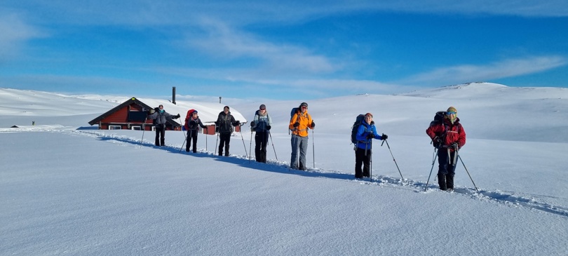 Vinterbilde med snø, hytte og en gruppe mennesker med ski på bena som står på rekke.