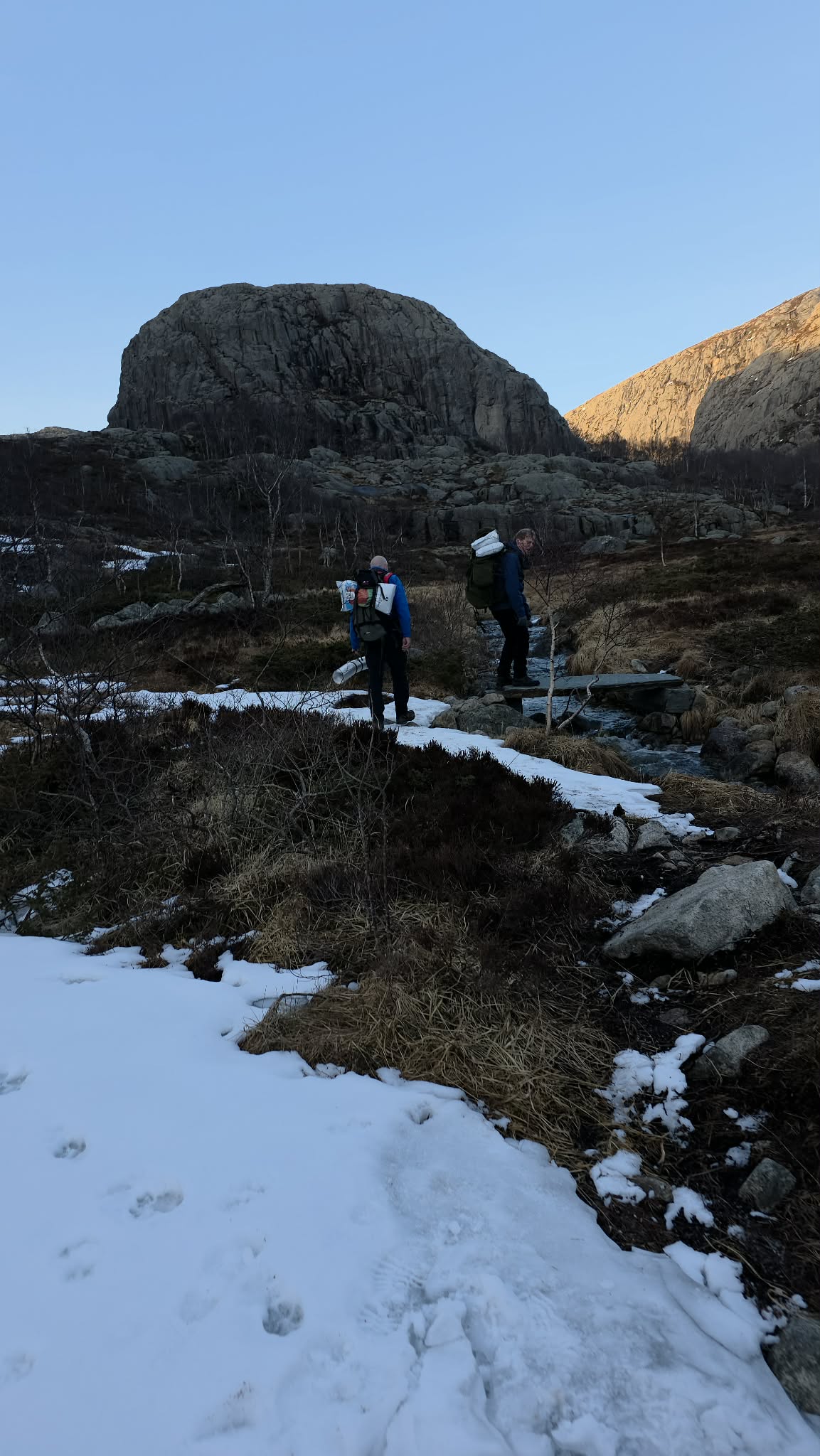 To personer med ryggsekk p&aring; tur i et landskap med fjell og noen sn&oslash;flekker p&aring; bakken.