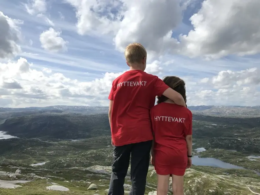 To barn, en gutt og en jente står og ser utover en landskap. De har røde t- skjorter på der det står hyttevakt.