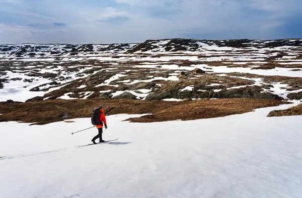 en person i rød jakke går på ski. Grønn hytte i horisonten. Det er litt snø men man ser det er vår 