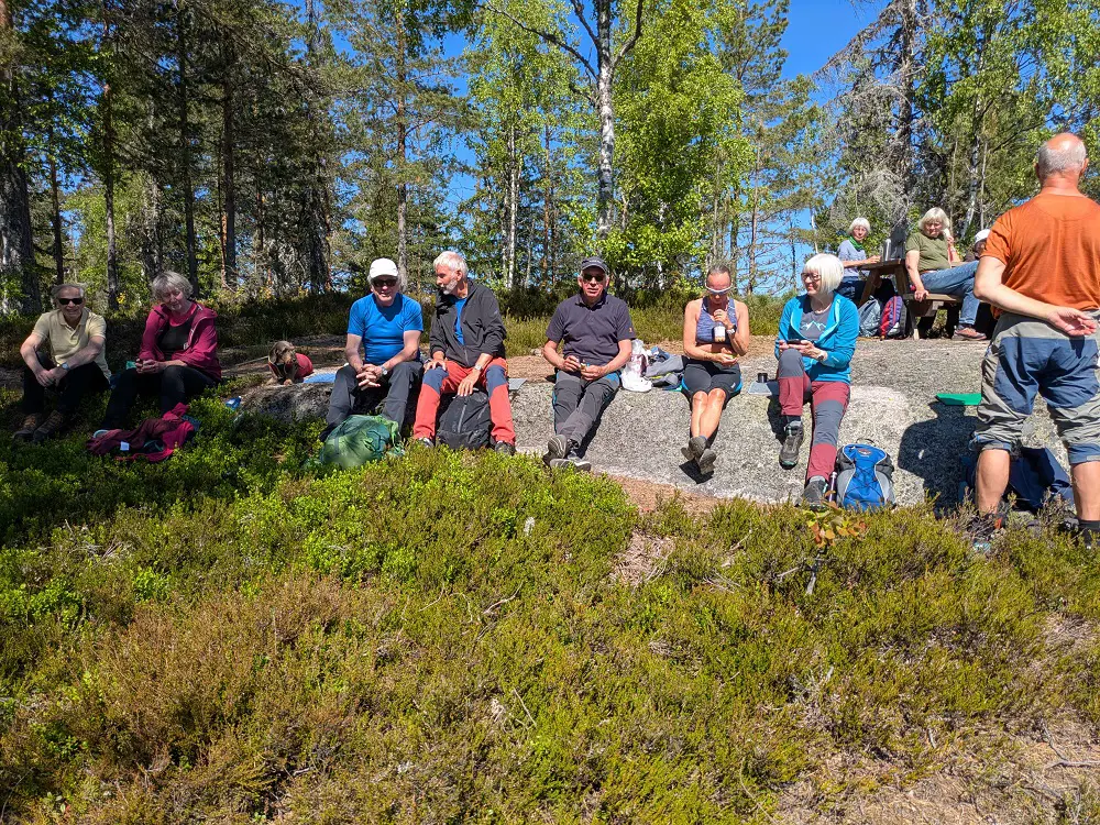 Personer som sitter på gresset. Det er blå himmel og bjørketrær i bakgrunnen. 
