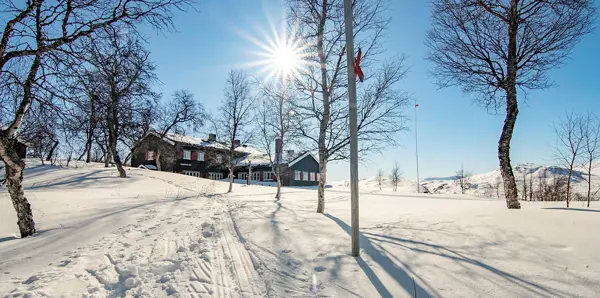 En brun L-formet to-etasjes hytte er synlig midt i bilder, der en ligger i en slak oppoverbakke, hvor hele terrenger rundt er dekket av snø. Spredte løvfrie trør står her og der. Solen stråler i en skyfri himmel. 