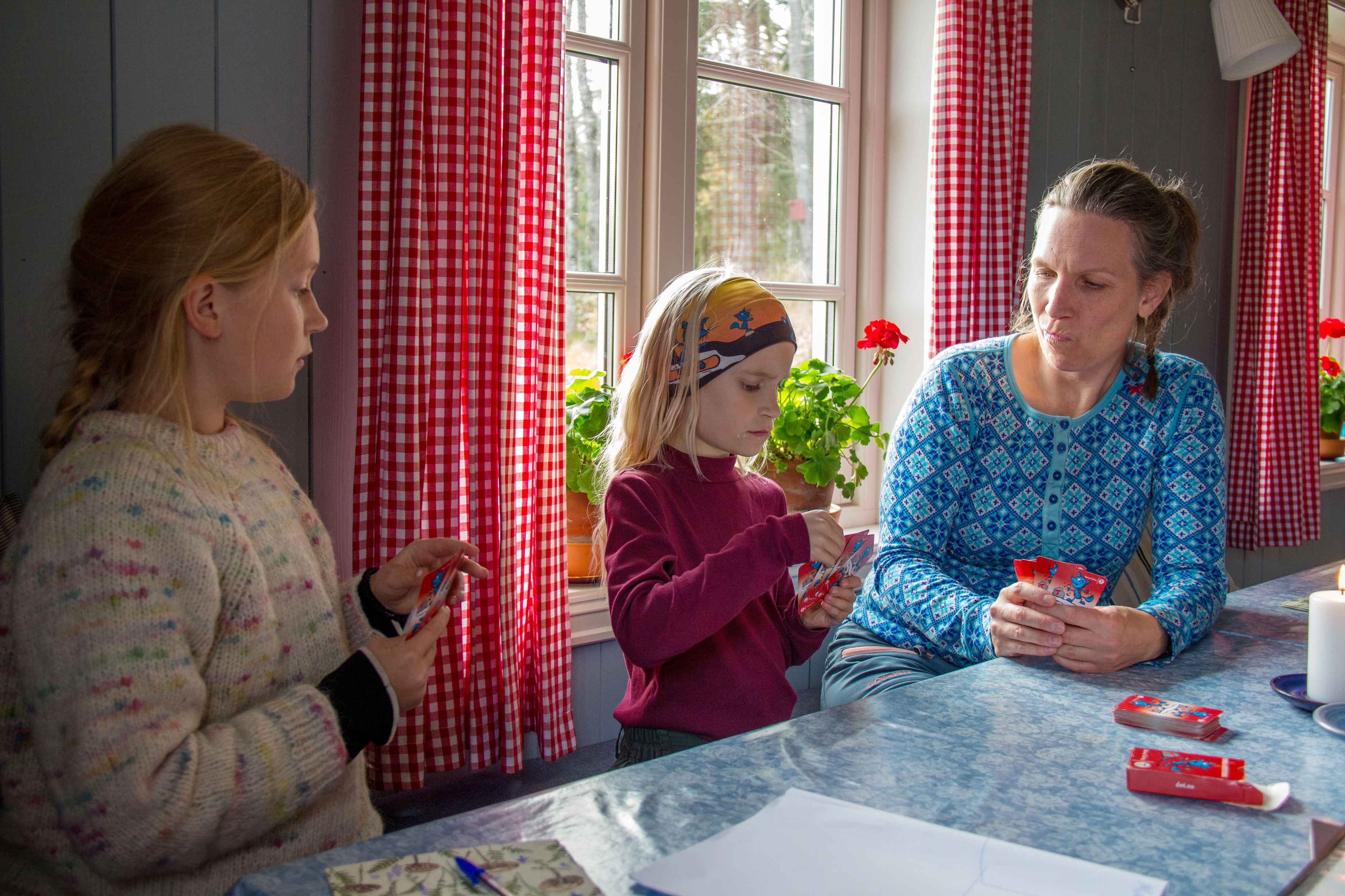 En dame og to jenter sitter inne ved et bord og spiller kort.