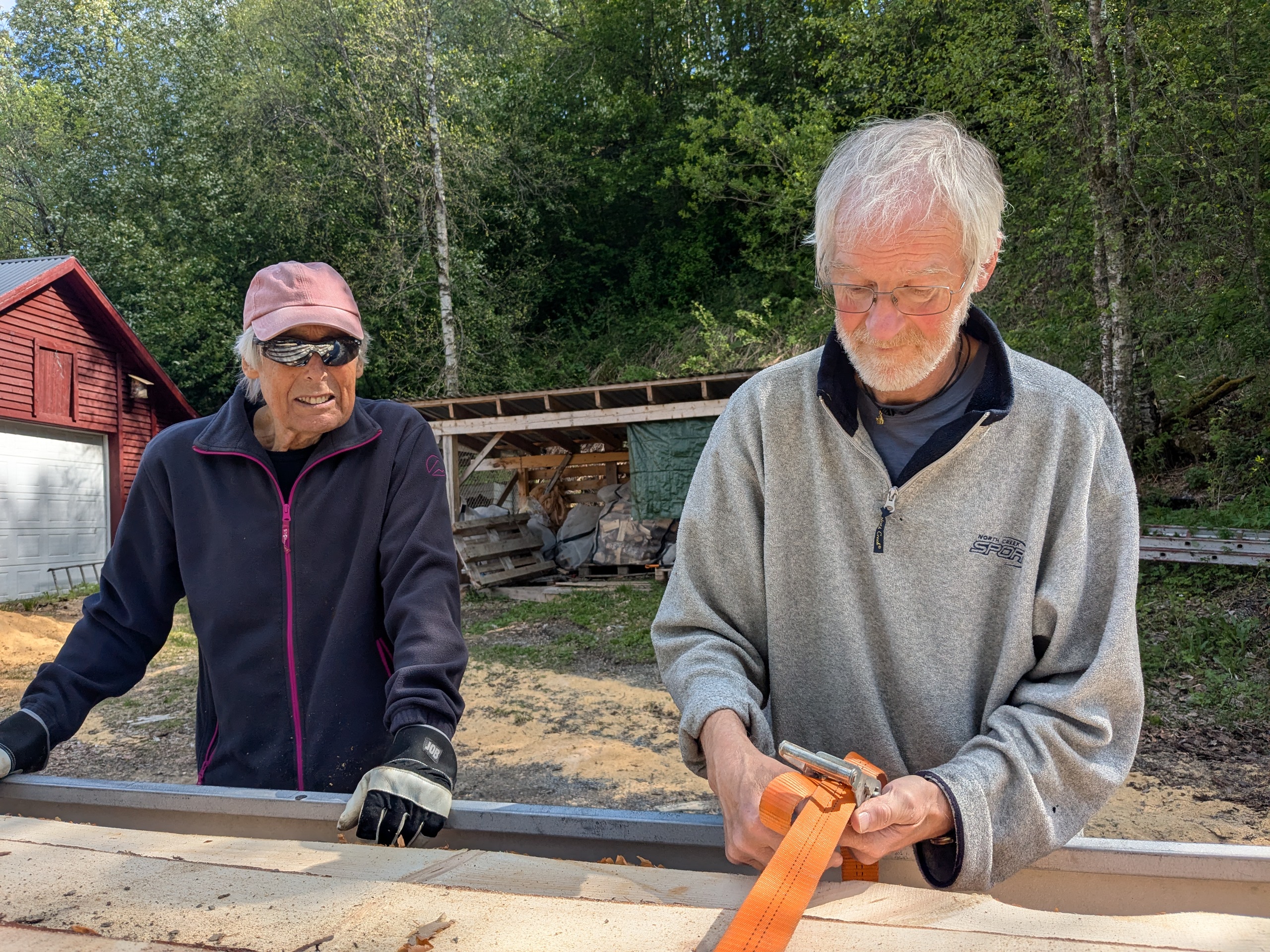 To eldre herrer ved tilhenger med planker. Bj&oslash;rkeskog i bakgrunnen