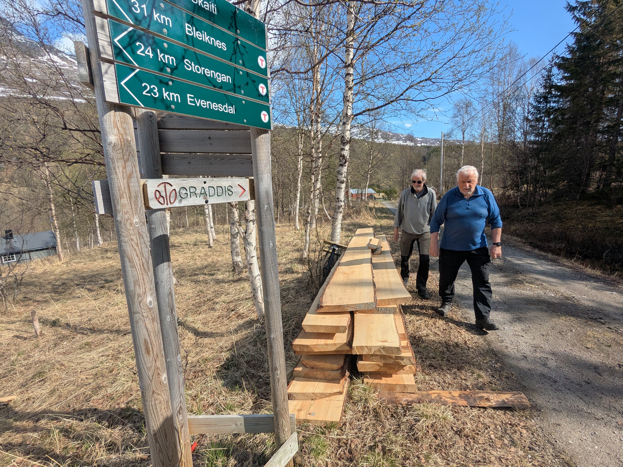 To eldre herrer b&aelig;rer planker. Skilt med retningsvisere ved siden av. 
