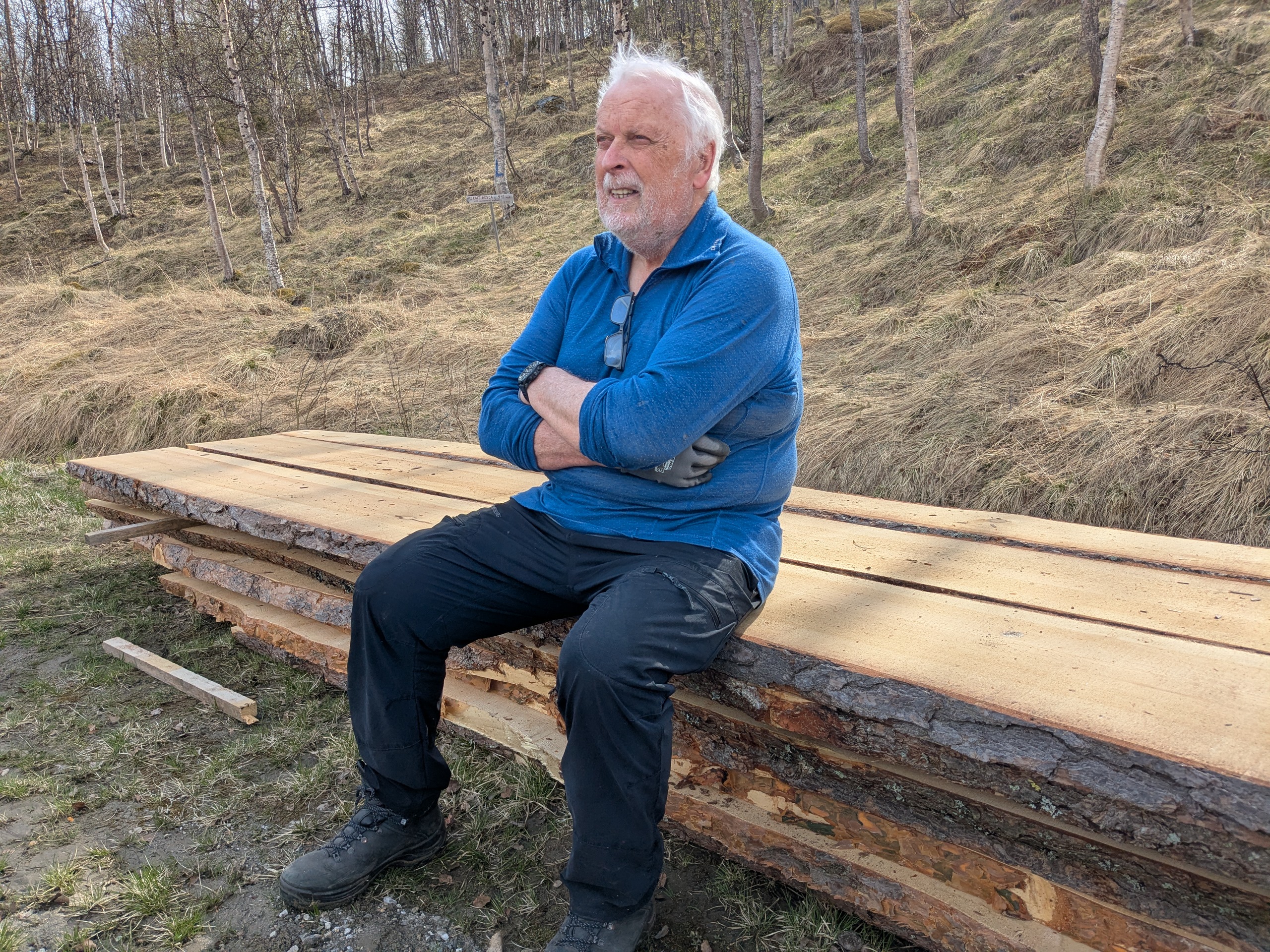 Eldre herre med solbriller hengende i skjorta tar seg en hvil p&aring; en plankehaug