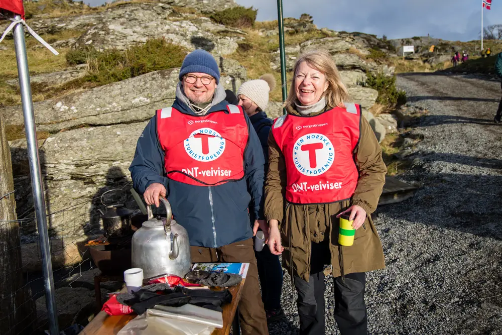 En mann og en kvinne med en rød vest med en stor T på brystet, står og smiler til kamera mens de koker kaffe utendørs.