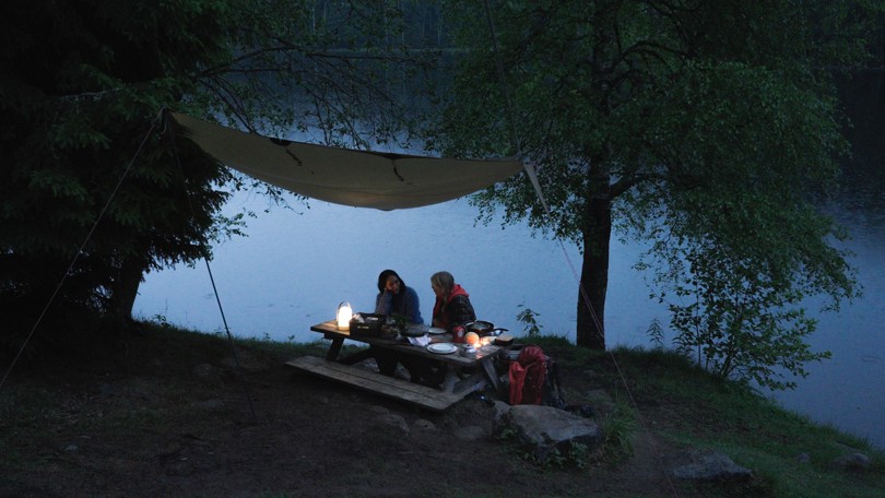 To personer sitter under en tarp ved et piknikbord ved bredden av en rolig innsjø, omringet av trær. Små lykter gir stemningsfull belysning i skumringen, og det ser ut som de nyter en utendørs middag eller samtale i den rolige, naturskjønne omgivelsen