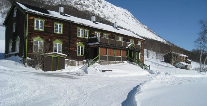 En brun to-etasjes hytte, med to-etasjesveranda foran, med snødekt landskap rundt og et fjell som ruver over bak hytta.