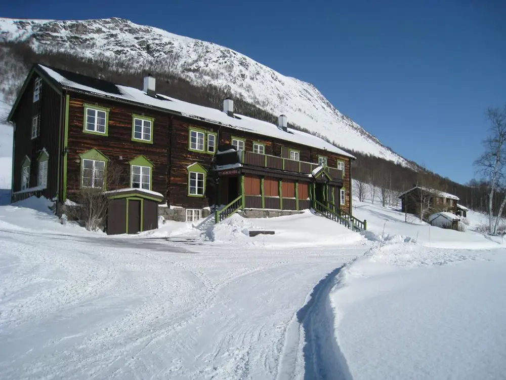 En brun to-etasjes hytte, med to-etasjesveranda foran, med snødekt landskap rundt og et fjell som ruver over bak hytta.