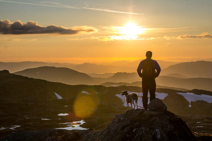 En mann og hunden hans står på et fjell og ser på solnedangen.