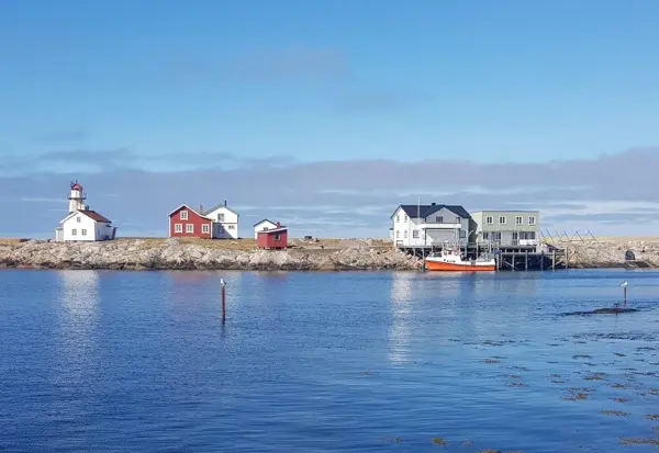 Syv små bygninger av treverk står spredt på en liten øy, to er rødmalte, resten er hvite. En ser ut som et lavt fyrtårn. Vannet mellom klippene og kamera er rolig.