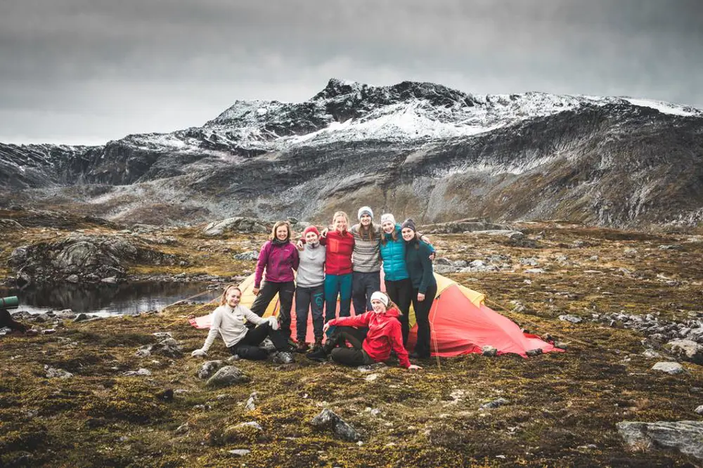 Åtte personer oppstilt foran et telt, to av dem sittende. Alle har på varme klær. Fjell i bakgrunnen.