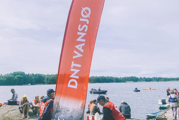 En gjeng med mennesker er samlet ved vannet, i front er det et DNT Vansjø beachflagg.