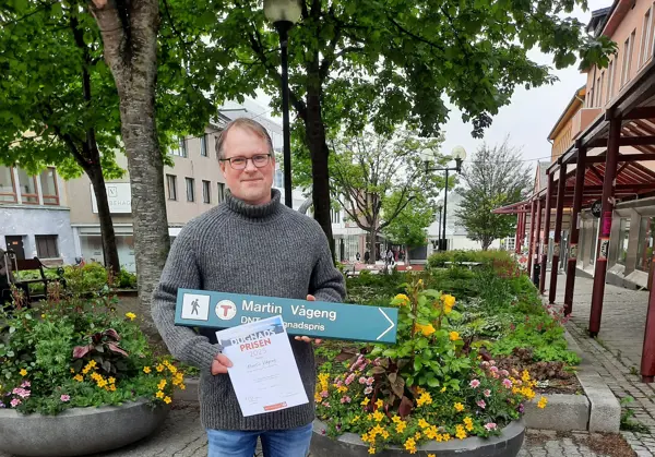 Mann foran trær og fine blomsterkrukker. Holder et grønt skilt og en diplom