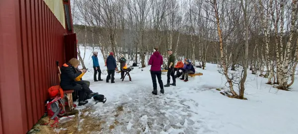 En gruppe mennesker med vinterklær står og sitter ved et rødfarget naust. Det er vinter og snø. 