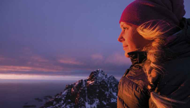 Nærbilde i profil av en kvinne i svart dunjakke og rød lue. I bakgrunnen skimtes et høyt, spisst og snøkledd fjell.