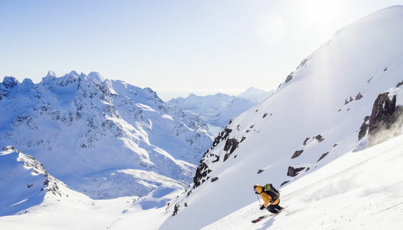 Skikjører i gul jakke kjører ned en bratt fjellside omgitt av spisse og alpine fjell.