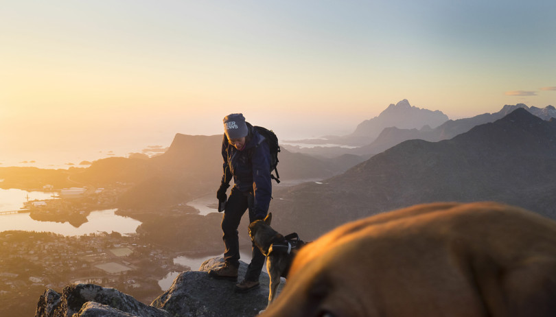 Person med hund står på toppen av et fjell i solnedgang.