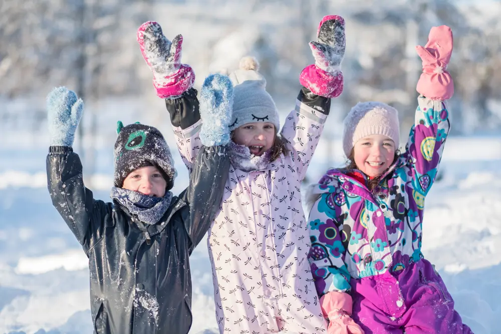 Tre glade barn i snøen som strekker henda i været. Foto.