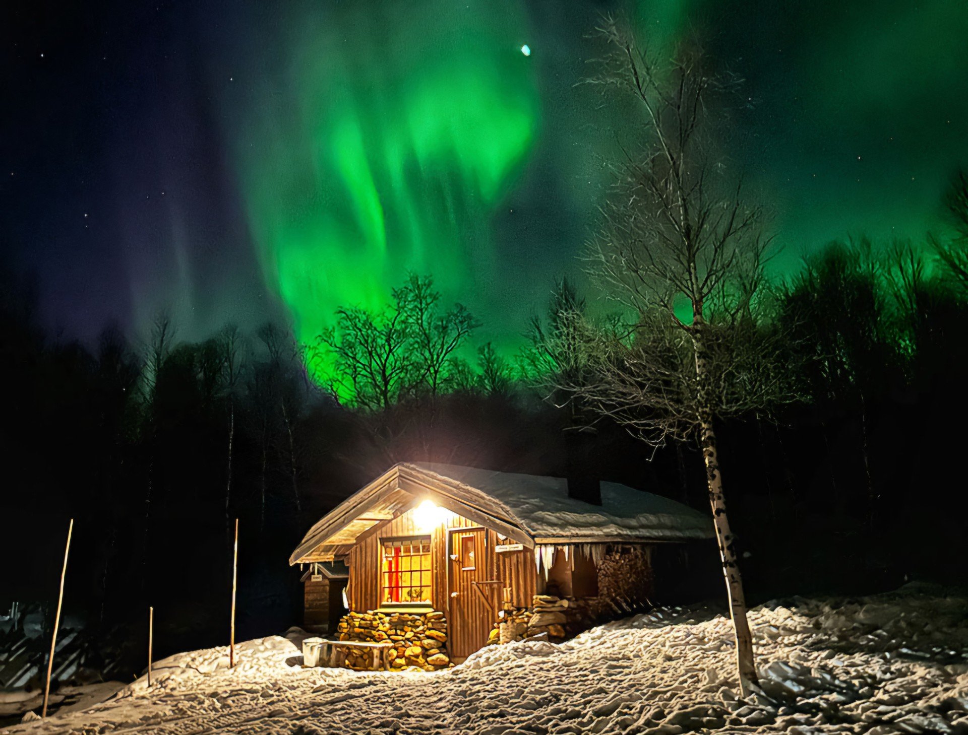Ei tømmerhytte med lys over trappa. Det er snø på bakken. Over hytta danser nordlyset grønt. 