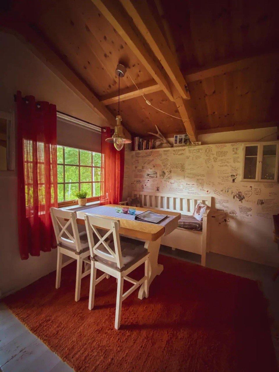 Ei hyttestue. En hvit benk og to hvite stoler på hver sin side av et bord. Rød gulvmatte og røde gardiner. 