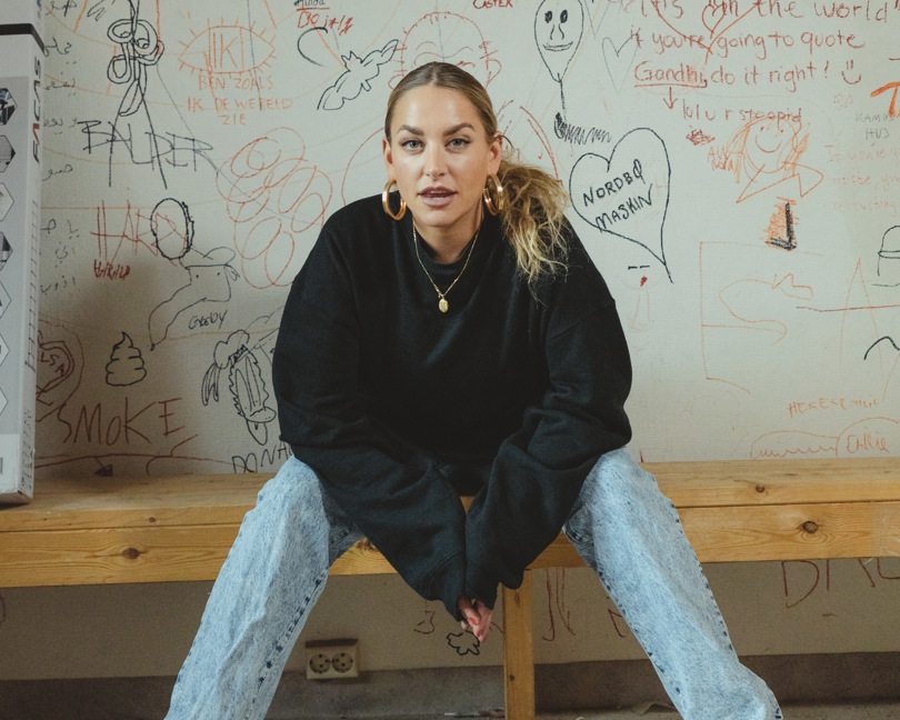Portrettbilde av en kvinnelig artist med langt lyst hår, sort genser og jeans. På veggen bak henne er det tegnet grafitti.