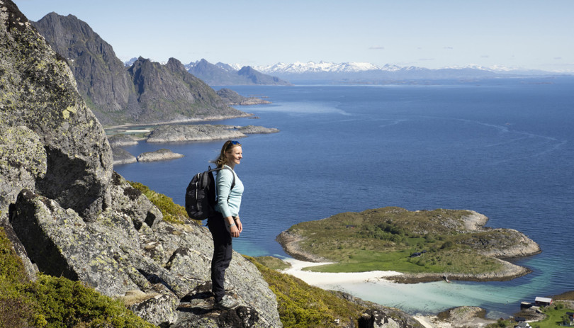 Kvinne med blå genser, svart bukse og ryggsekk står i en fjellside og ser ut over høye fjell og hav.