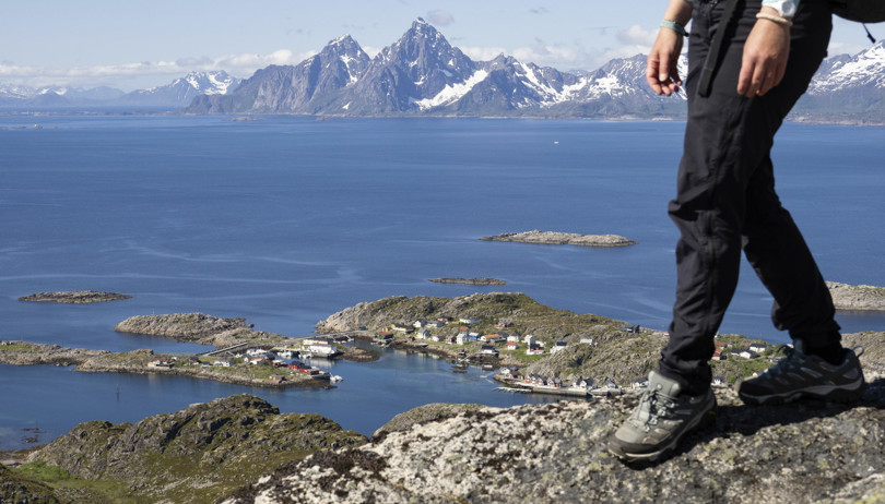 Person (vi ser bare personen fra hoften og ned) går over en stein på et fjell. Nedenfor fjellet ligger et lite fiskevær, og i bakgrunnen er det hav og høye fjell.