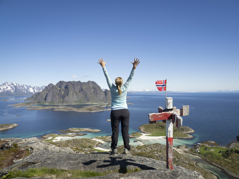 Person i blå genser og svart bukse står med ryggen til og strekker armene i været på toppen av et fjell omgitt av hav, øyer og fjell. Ved siden av henne står det en liten stolpe med et norsk flagg på toppen.