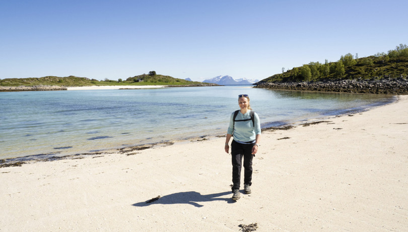 Person i blå genser og svart bukse går over en kritthvit strand. I bakgrunnen skimtes en annen strand og høye fjell. 