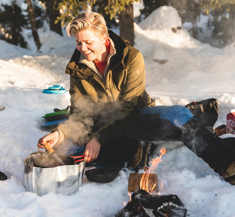 Person sitter ved et bål i snøen og rører rundt i en kjele med mat. 