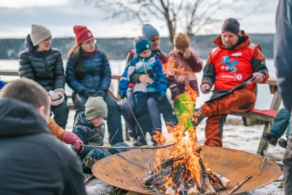 Voksne og barn som griller pølser på bålpanne. Foto.