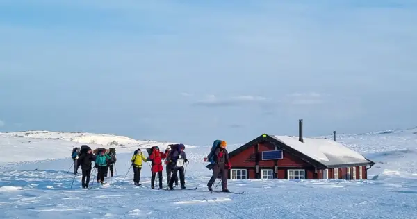 En gruppe mennesker med vinterbekledning, sekk på ryggen og ski på bena på vei fra ei brunrød hytte. Det er vinter og snø. 