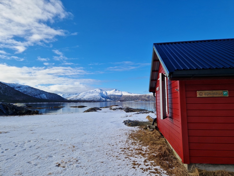 En rød hyttevegg. Det er snø på bilde. Bak ser vi havet og noen høye, snøkledte fjell. 
