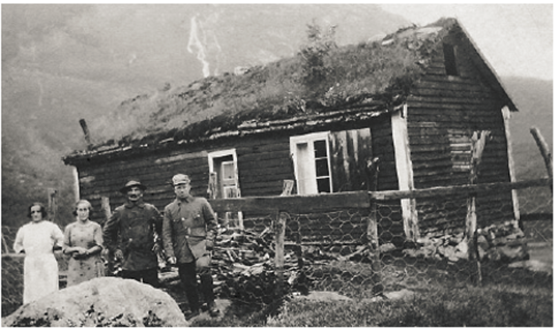 Svart-hvitt-bilde av fire mennesker foran gammel hytte. Foto.