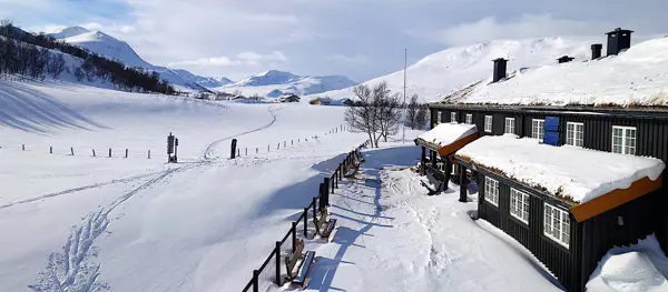 Langsiden av en brun to-estasjes hytte tar opp høyre kant av bildet, med et gjærde foran som skillet det lille tunet foran hytta fra det store og snødekte landskapet som strekker seg utover i resten av bildet. Helt hvit-dekte fjell i bakgrunnen.
