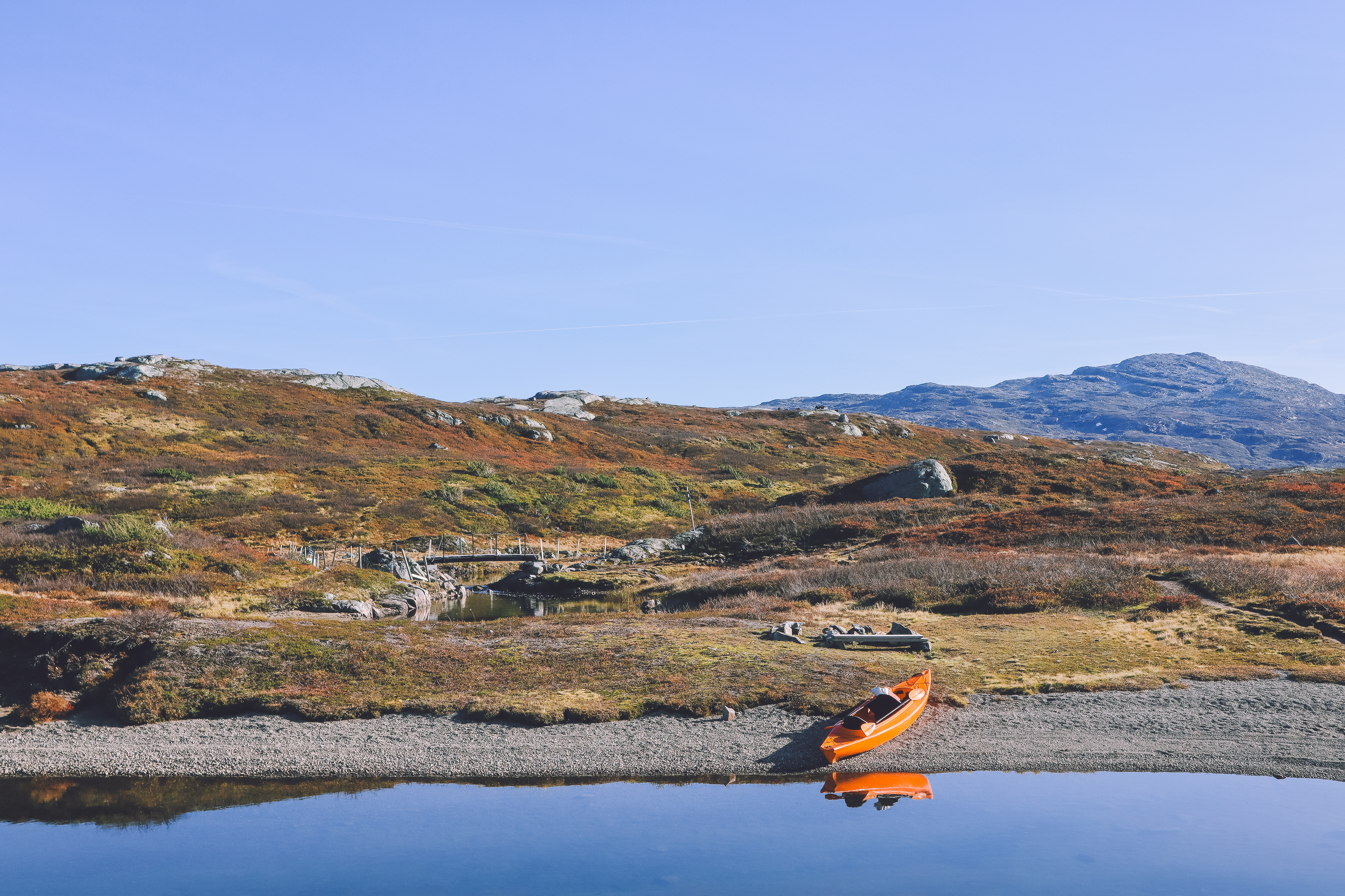 En orange kajak p&aring; land ved et speilblankt vann, h&oslash;stfargede fjell i bakgrunnen.