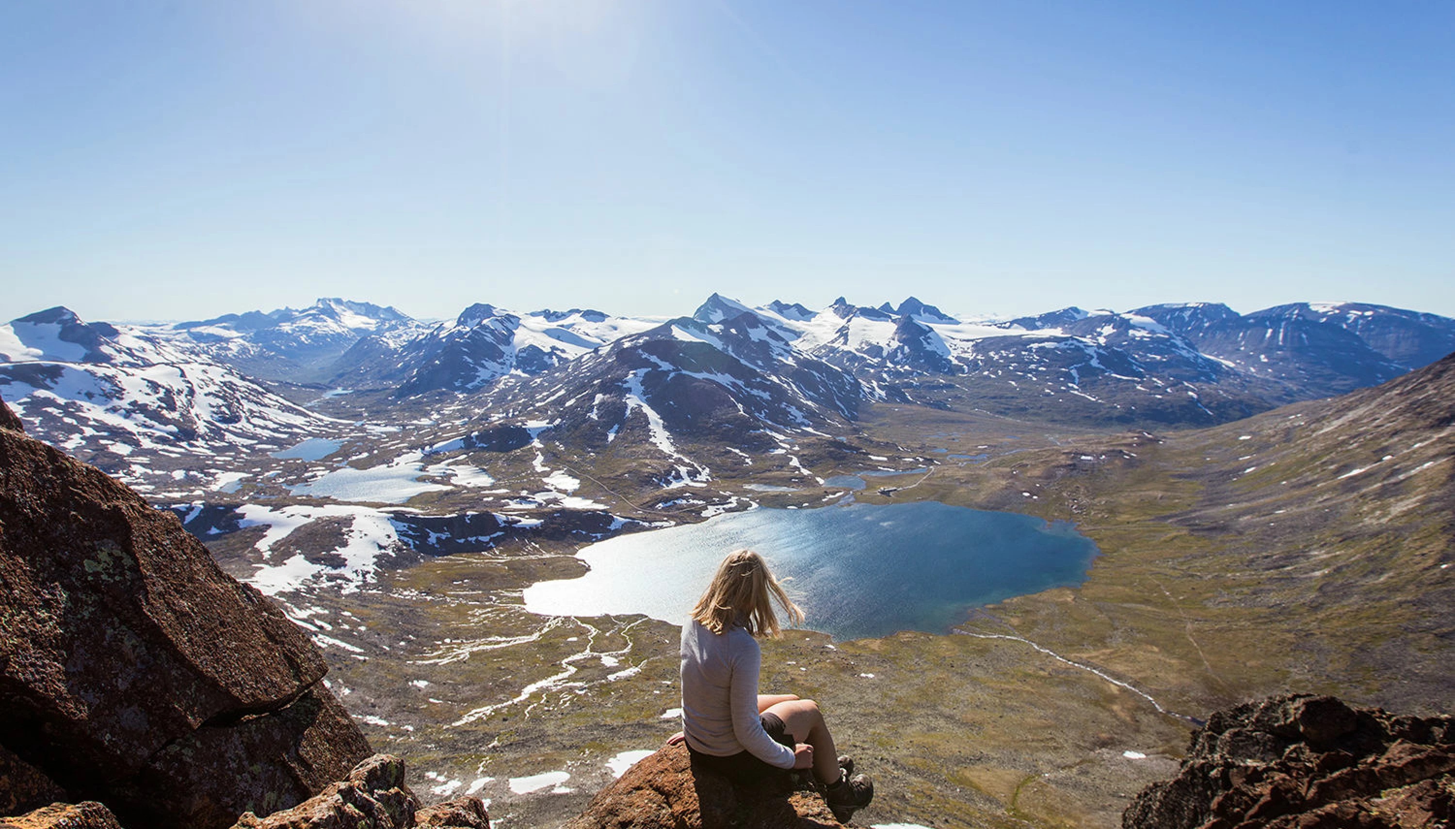 Kvinne sitter ytterst på en stein på toppen av et fjell og skuer utover fjellheimen.